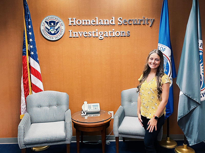 Woman standing in front of flags and wall sign that says Homeland Security Investigations