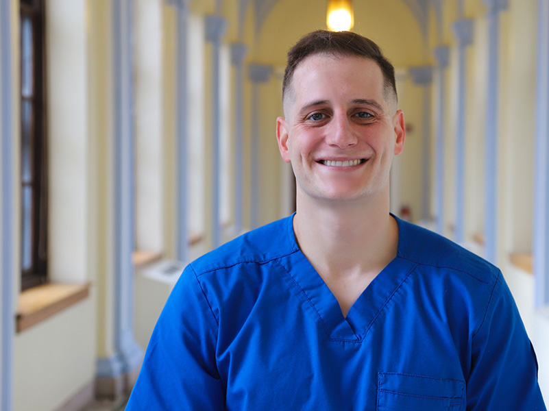 Man in hallway, wearing blue scrubs.