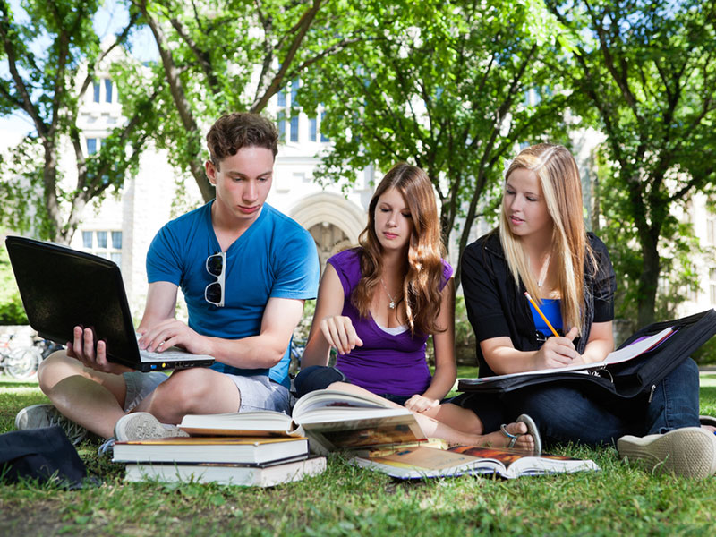 Three college students sitting on grass with laptop and books.