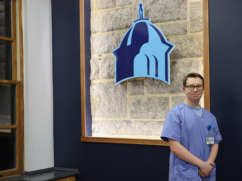Student in scrubs standing in from of Immaculata logo
