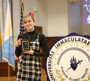Young woman holding plaque in front of large college seal