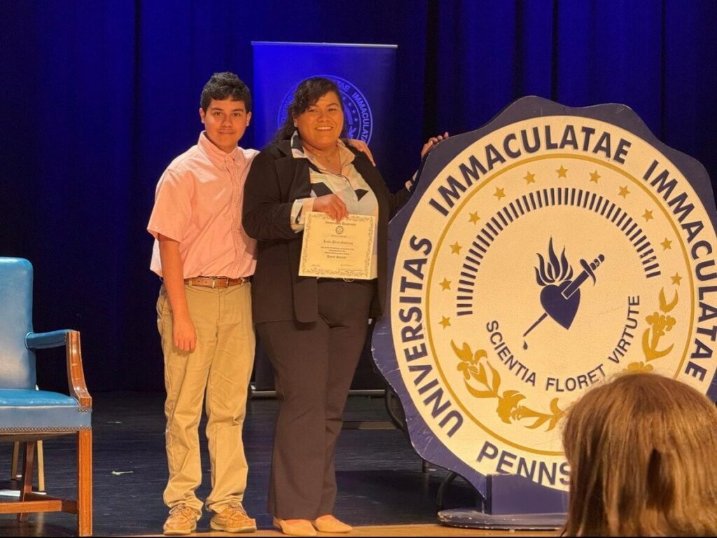 A woman stands on a stage at Immaculata University holding a certificate, smiling as a young boy stands beside her with his arm around her. They pose next to a large Immaculata University seal against a blue-lit backdrop, suggesting a formal academic recognition or awards ceremony.