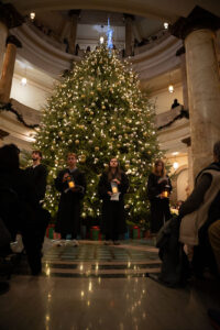 People in robes singing and holding lanterns in front of Christmas tree