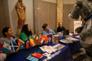 Mascot talks to group of people behind table. Flags of different countries are on the table
