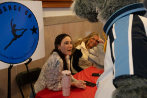 Two women behind table talk to mascot