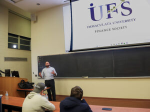 IU alum, Dan Klodarska, stands in front of a chalkboard, speaking to students at the inaugural IU Finance Society meeting. Two students are also seen sitting in the foreground.