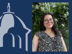 A headshot, color photograph taken during the daytime shows Immaculata graduate student, Jaylynn Carey ,with light-medium skin and dark hair smiling. She has shoulder-length, dark, straight, or slightly wavy hair parted slightly to her right, and wears round black-framed eyeglasses and small hoop earrings in both ears. She wears a silver-colored chain with a small J-shaped pendant and a leopard-print short-sleeved top that is black and shades of beige and cream with ruffled sleeves and a ruffled collar that is tied.