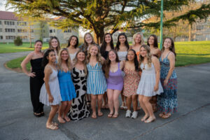 Group of smiling young women in sundresses posing together on a sunny campus lawn, with a tree and buildings in the background.