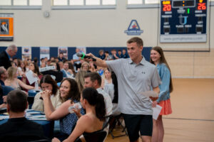 Male student emcee in a gym by a crowded banquet-style event, holding a microphone and addressing the audience while a smiling girl stands behind him holding papers