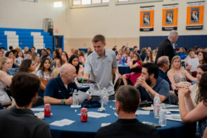 Young man in a gray polo stands and speaks into a microphone at a round banquet table, with students watching in a gymnasium.