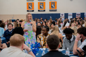 A girl in a colorful floral dress speaks into a microphone while reading from a sheet to a large seated student audience at a school event in a gymnasium.