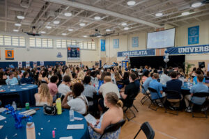 Busy school gym filled with round tables and attendees facing a stage with a blue banner reading Immaculata University.