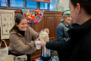 Woman pours tea from a teapot into a cup handed to a guest at a community event, smiling warmly at the moment of service.