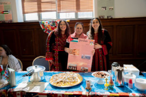 Three women in colorful traditional outfits hold a Jordan information poster at a cultural booth with food on the table behind them.
