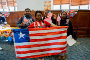 Four diverse children pose indoors, holding a large Puerto Rico flag and waving small American flags at a cultural event.