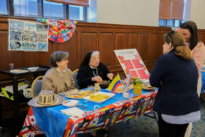 A cultural information table with international flags, two women and a nun talking to a standing visitor, snacks and a Colombia brochure on the table.