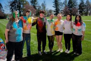 Group of seven friends in a park, arms around each other, covered in colorful powder and smiling at the camera.