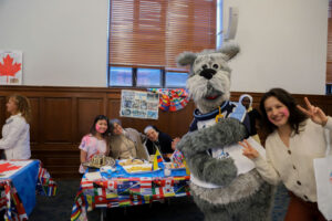Costumed gray dog mascot and smiling woman pose beside a table decorated with international flags at a multicultural event.