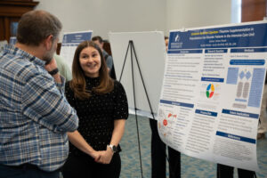 Man in a plaid shirt converses with a smiling woman beside a science poster at a conference.