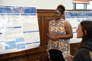 Woman in a patterned sleeveless top explains a research poster to an audience in a room; another person seated nearby with a drink listens.