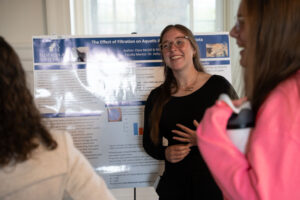 Woman presenting a research poster at a science fair or conference, smiling and gesturing beside the poster boards.