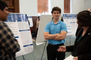 Person in a blue polo stands with arms crossed next to a science poster at a conference poster session.