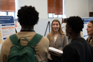 Woman in a gray blazer presenting a poster at a conference, speaking to a small group of attendees in a room lined with posters.