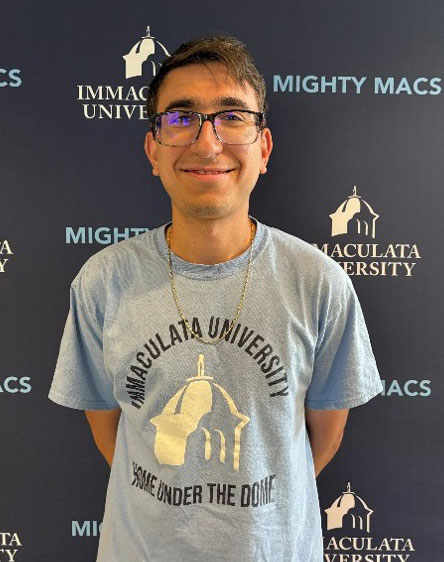 Ethan Dwyer - Immaculata University Smiling man with glasses in a light blue Immaculata University t‑shirt, posing in front of a backdrop bearing Immaculata University and Mighty Macs logos.