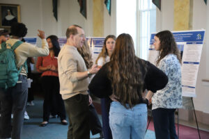 Group of students discussing a research poster at a campus poster session, with boards and people listening in the background.