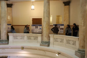 People examine exhibit posters along a marble balcony in a classical interior with tall columns and warm lighting.