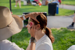 Face painter applies a pink flower design to a woman's cheek outdoors in a park scene.