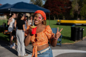 Smiling woman with face paint and an orange top holding a red drink and making a peace sign at an outdoor party.