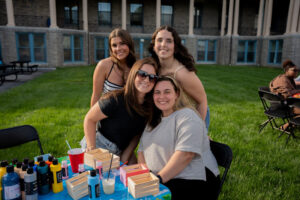 Four smiling young women posing together at an outdoor craft table with paint bottles and wooden boxes visible on the table.",