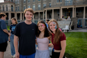 Three friends pose outdoors for a photo, two women and a man, with colorful face paint on their cheeks, smiling at the camera.