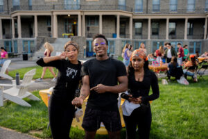 Three friends pose for a photo at an outdoor campus gathering on a grassy lawn with people socializing in the background.