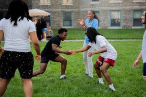 Group of friends playing a handshake-style game on a grassy campus lawn, cheering and smiling.
