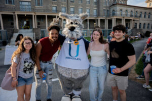 Five students pose with a gray wolf mascot wearing an Immaculata banner outside a stone campus building.