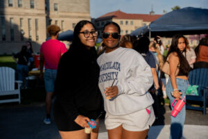Two women posing at an outdoor campus event; one in a gray MACULATA UNIVERSITY sweatshirt, the other in black, both smiling.