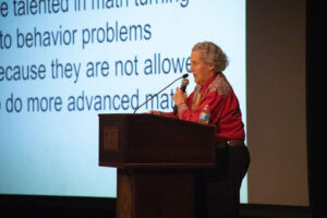 Older woman in a red blouse speaks at a wooden podium with a microphone, in front of a large text slide.