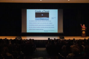 Presenter giving a talk on a stage with a projected slide titled 'Create a Planet' showing bullet points about students and planning, with a speaker at a podium on the right and a dark theater filled with an audience.