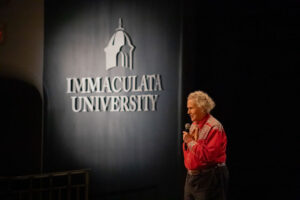 Woman with microphone speaking on a stage beside a banner that reads Immaculata University.