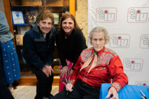 Three women lean in beside an elderly woman in a red shirt seated at a blue-covered table, with a logo backdrop behind them.
