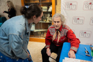 Young woman in a denim jacket talks with an elderly woman seated at a blue table during a community event, backdrop with organization logos behind them.