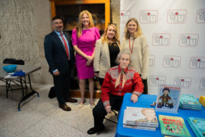 Five adults posing for a group photo indoors at an information table with brochures and a branded backdrop.