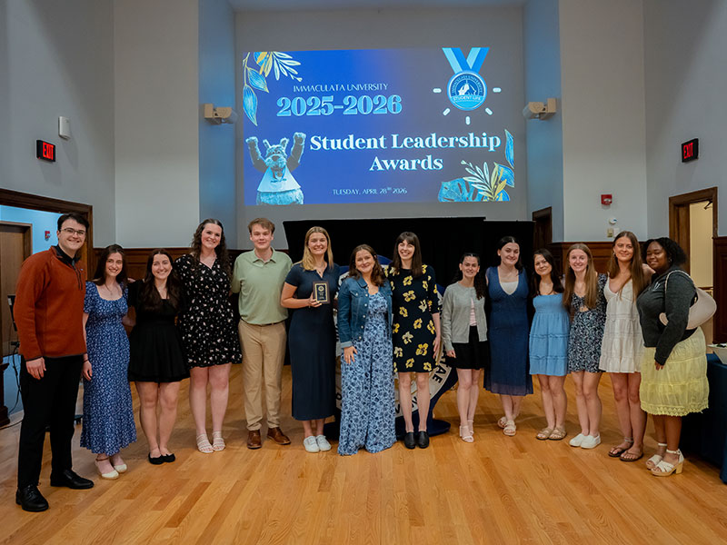 Group of students in semi-formal outfits posing on a stage after receiving leadership awards, with a blue screen reading 'Student Leadership Awards' in the background (Immaculata University).