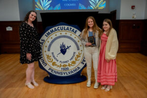Three young women pose beside a large circular seal reading Universitas Immaculatatae Pennsylvania; one holds a plaque at an awards event.