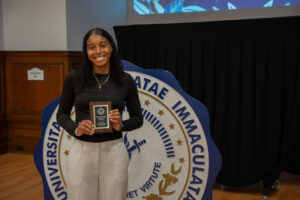 Woman in black top and white pants holding a plaque at a recognition event, standing beside a large university seal backdrop.