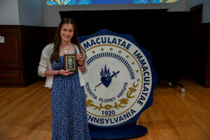 Smiling young woman in a blue floral dress holds an award plaque beside a large circular seal on a stage.