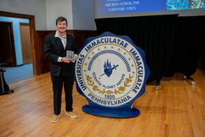 Man in a dark suit holds a small award while standing beside a large Immaculata University, Pennsylvania seal on a wooden floor.