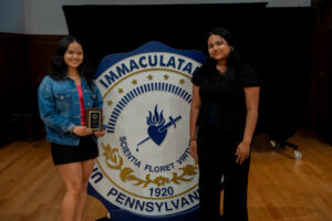 Two women pose beside a large Immaculata University seal; the woman on the left holds a small plaque.